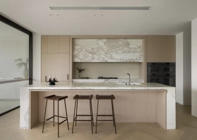 Minimalist kitchen with light wood cabinets, marble countertops and backsplash, built-in appliances, and two modern wooden stools at the island. A large window allows natural light into the clean, open space.